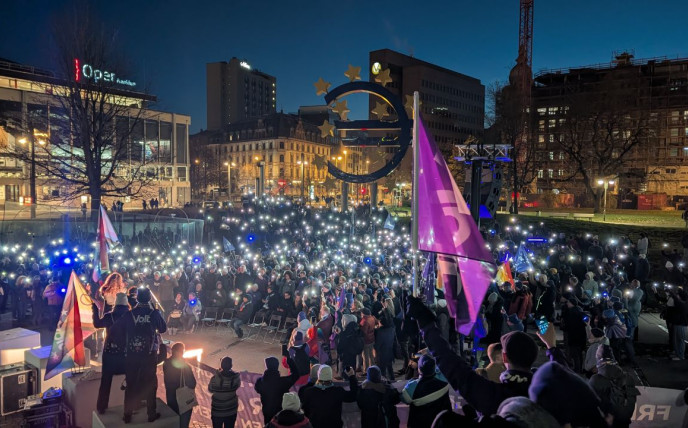 Eine nächtliche Aufnahme einer grossen Kundgebung auf einem Platz in einer Stadt, wahrscheinlich in Deutschland. Hunderte von Menschen haben sich versammelt und halten ihre leuchtenden Smartphone-Taschenlampen in die Luft, was einen beeindruckenden Anblick erzeugt. Im Hintergrund, zwischen den Stadtgebäuden, dominiert eine grosse, beleuchtete Skulptur der zwölf goldenen Europasterne in einem Kreis. Im Vordergrund, auf einer erhöhten Plattform (Bühne), stehen mehrere Personen, eine davon trägt eine Weste mit dem 