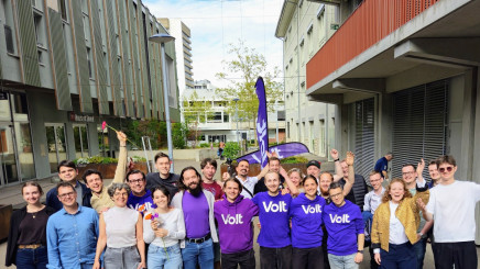 Eine Gruppe von etwa 20 gut gelaunten Menschen posiert im Freien auf einem asphaltierten Platz vor modernen Gebäuden. Mehrere Personen in der Mitte tragen violette T-Shirts oder Hoodies mit dem weissen Aufdruck «Volt». Einige Personen halten Blumen hoch oder haben die Arme jubelnd in die Luft gestreckt. Im Hintergrund ist eine violette Volt-Fahne zu sehen. Das Bild vermittelt eine positive, gemeinschaftliche Atmosphäre.